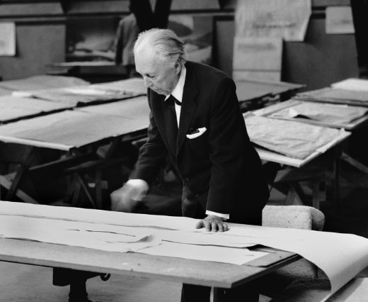 Archival photography of Frank Lloyd Wright, looking over architectural plans in his drafting studio.