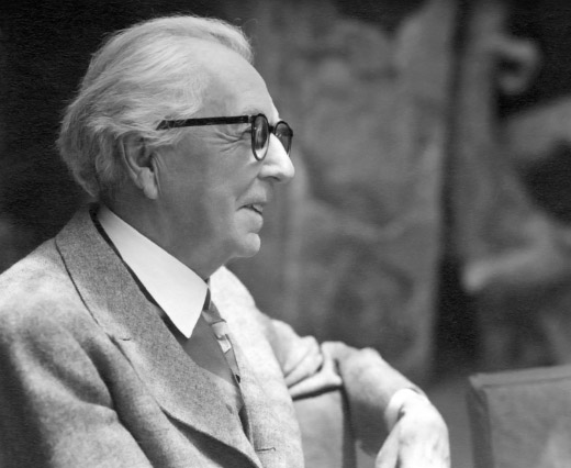 Archival photography of Frank Lloyd Wright, wearing glasses and smiling as he turns away from the camera.